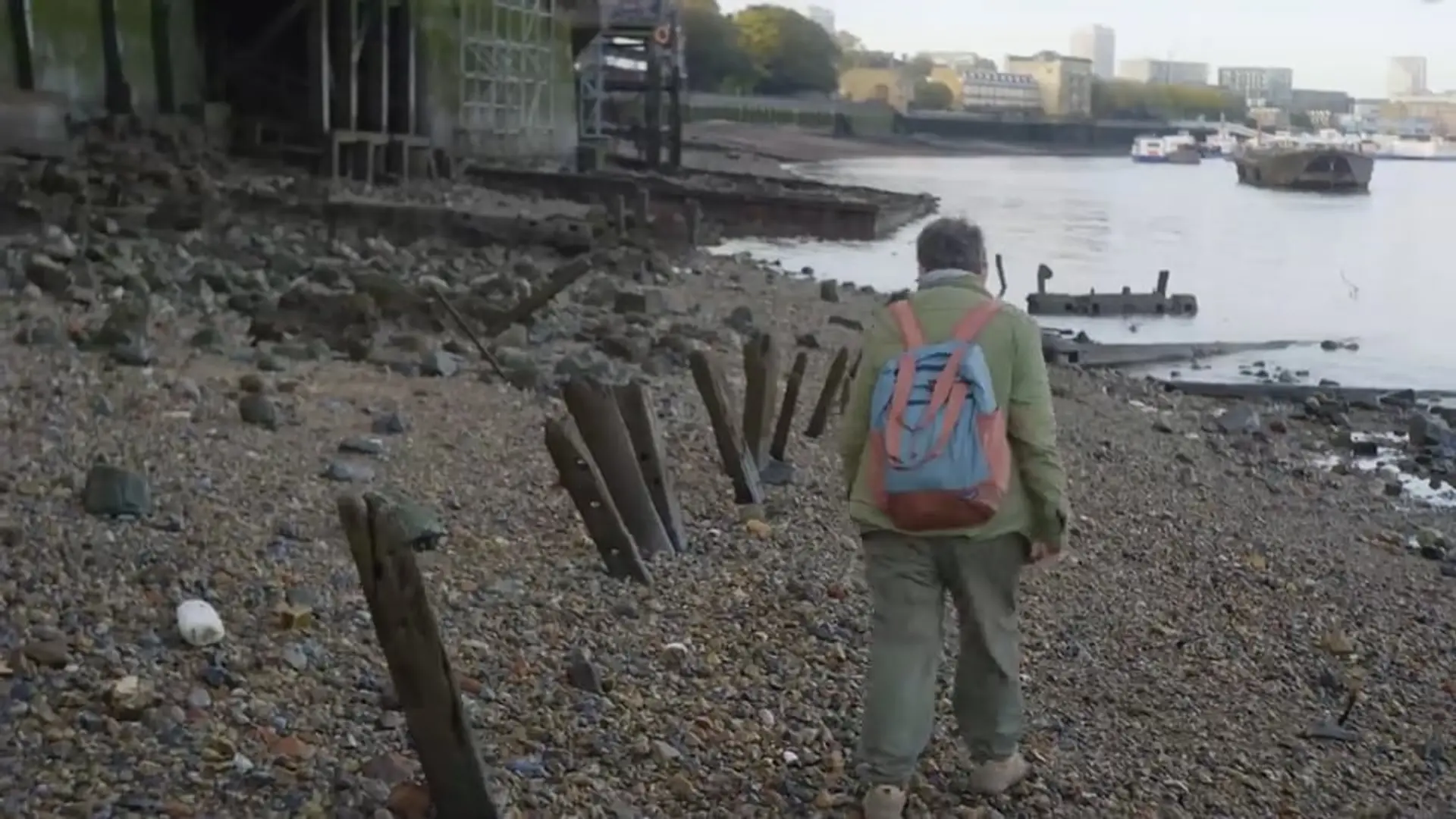 A person with a backpack walks along the rocky Thames riverbank, surrounded by remnants of structures.