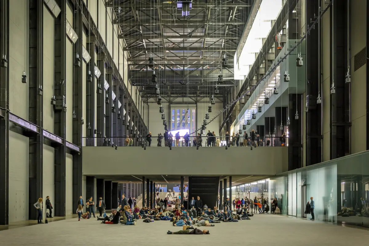 Tate Modern's Turbine Hall, a high-ceilinged gallery space, filled with individuals in various positions such as standing, sitting, leaning against walls, and lying on the floor.