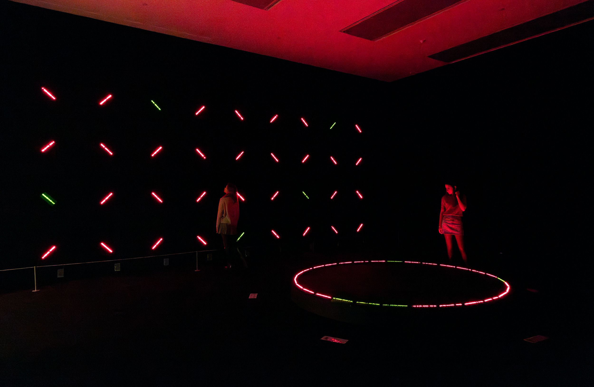 A dark room illuminated by a grid of diagonal red and green LED lights on the wall and a glowing circular platform on the ...