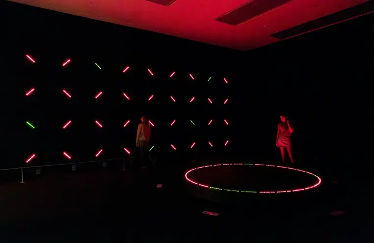 A dark room illuminated by a grid of diagonal red and green LED lights on the wall and a glowing circular platform on the ...