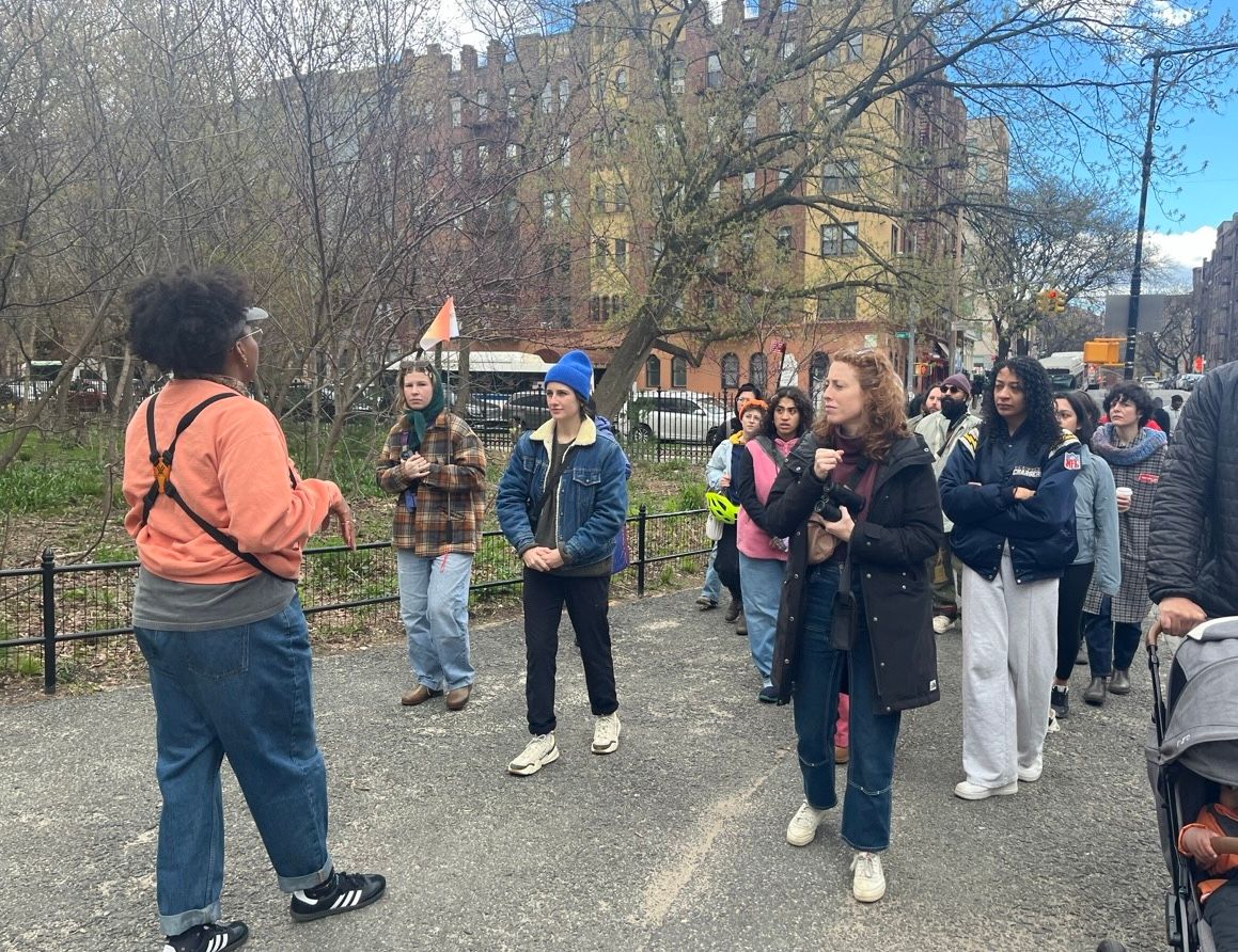 A person leading a group of people on a bird walk in an urban park, with participants attentively listening and some holding binoculars.