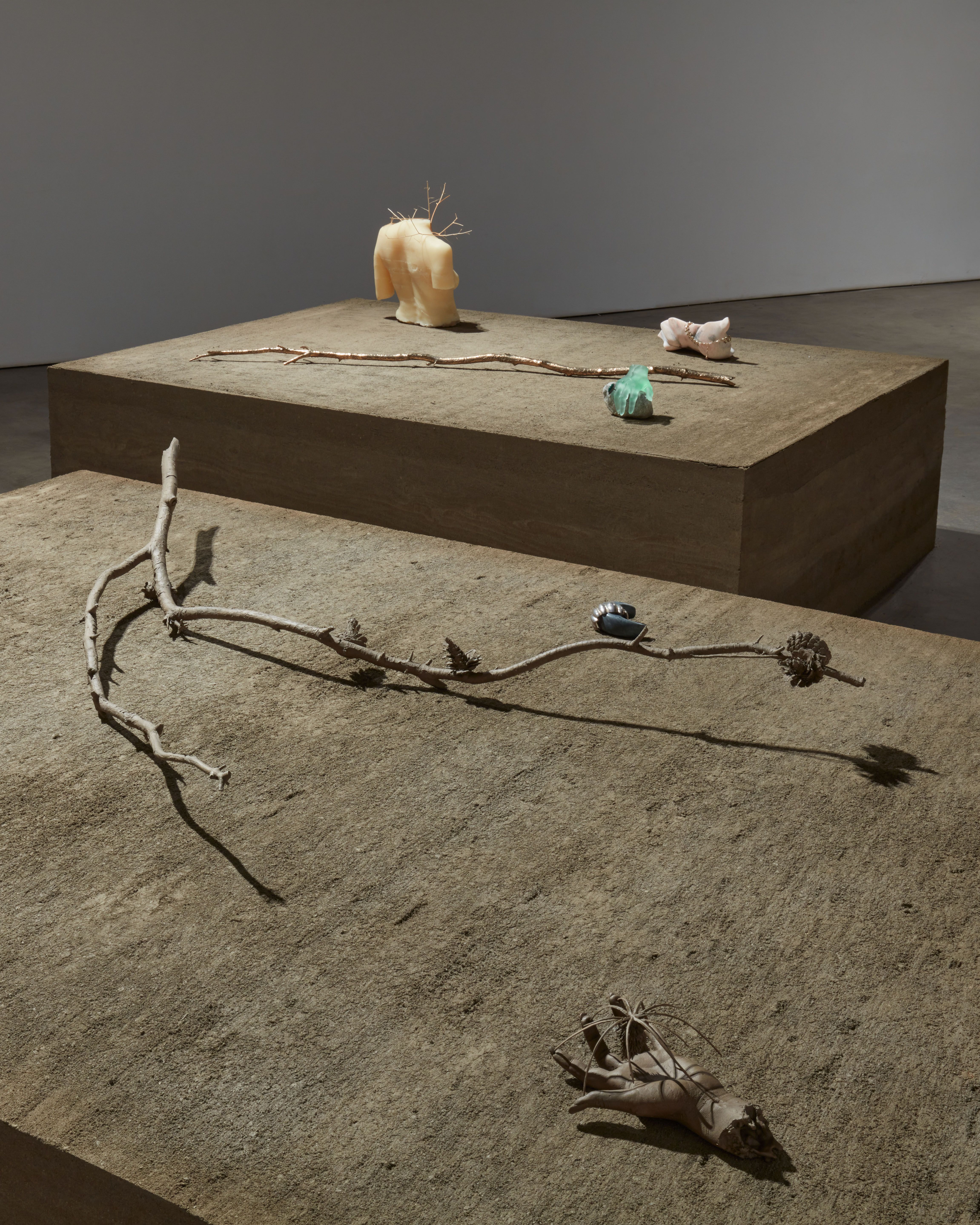 An art installation featuring a rectangular wooden table displaying twigs, a headless upper body statue, a sculpted hand, and an aqua colored rock.