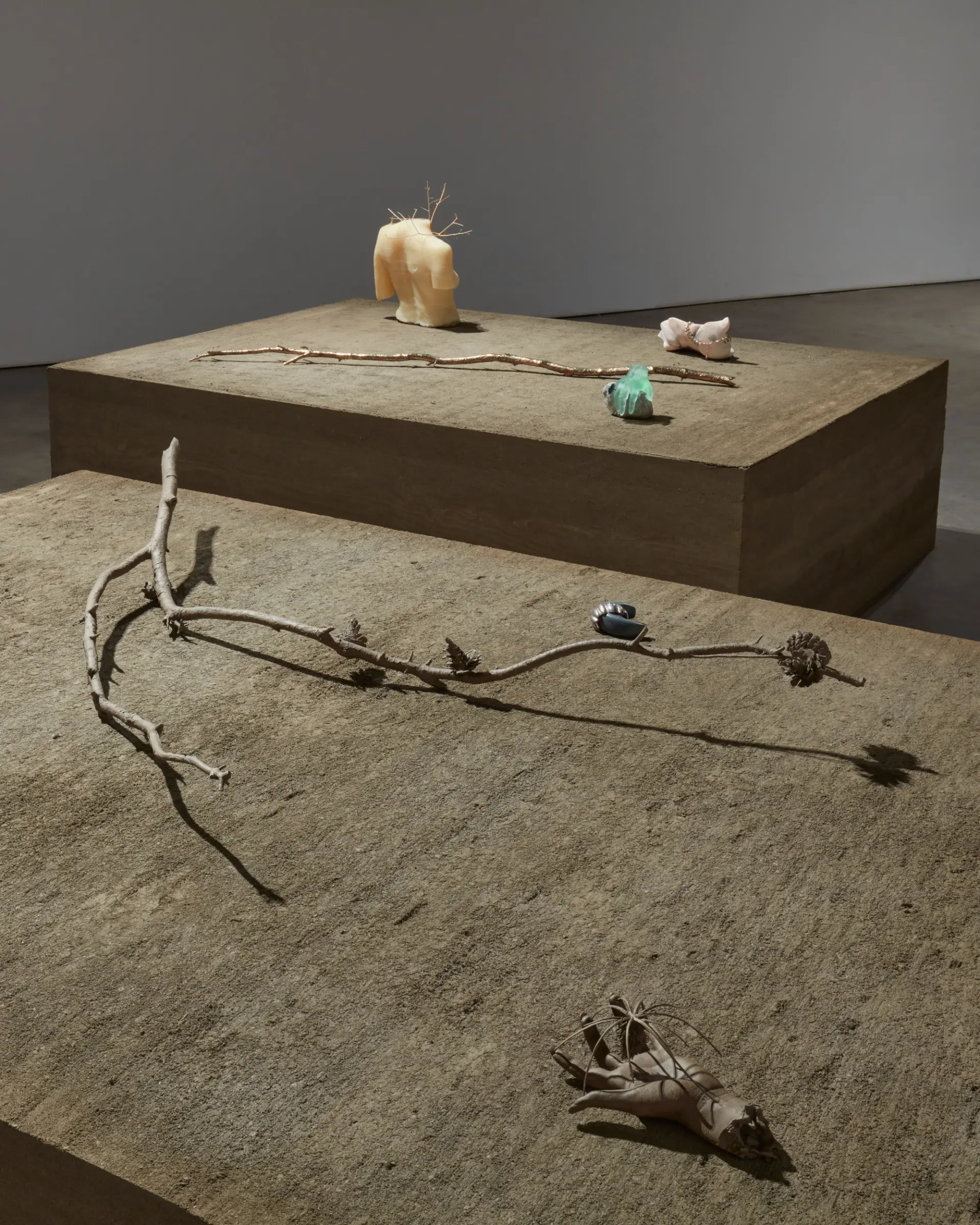 An art installation featuring a rectangular wooden table displaying twigs, a headless upper body statue, a sculpted hand, and an aqua colored rock.