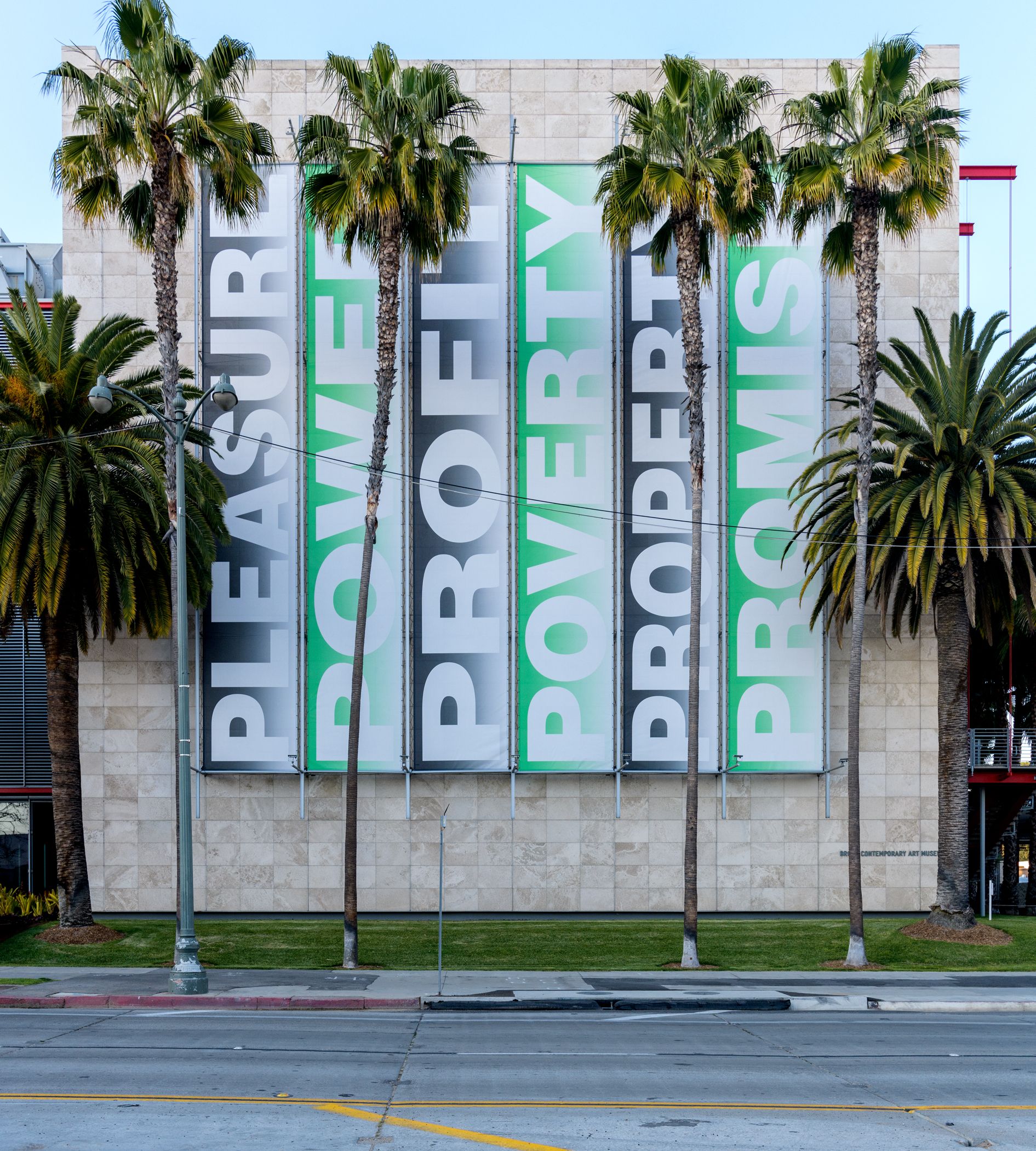 A building, nestling among tall palm trees, features a large wall advertisement with the text "Pleasure, Power, Profit, Po...