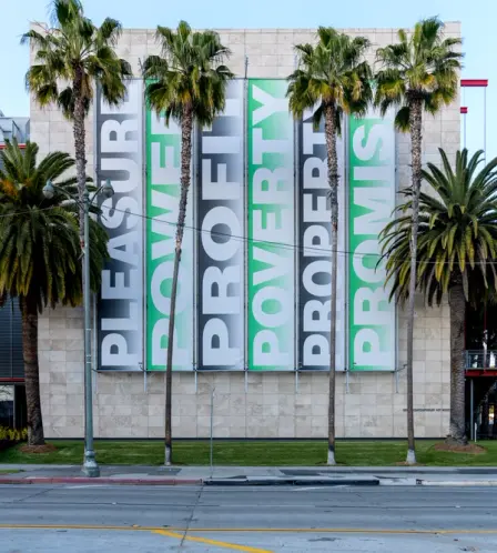 A building, nestling among tall palm trees, features a large wall advertisement with the text "Pleasure, Power, Profit, Po...