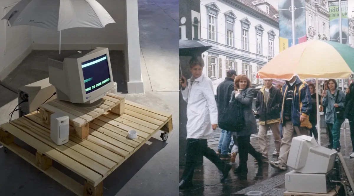 An outdoor art installation featuring an old desktop computer placed on a wooden pallet with wheels, topped with an umbrella. The second image shows the same installation on a city sidewalk, with people passing by under umbrellas and city buildings in the background.