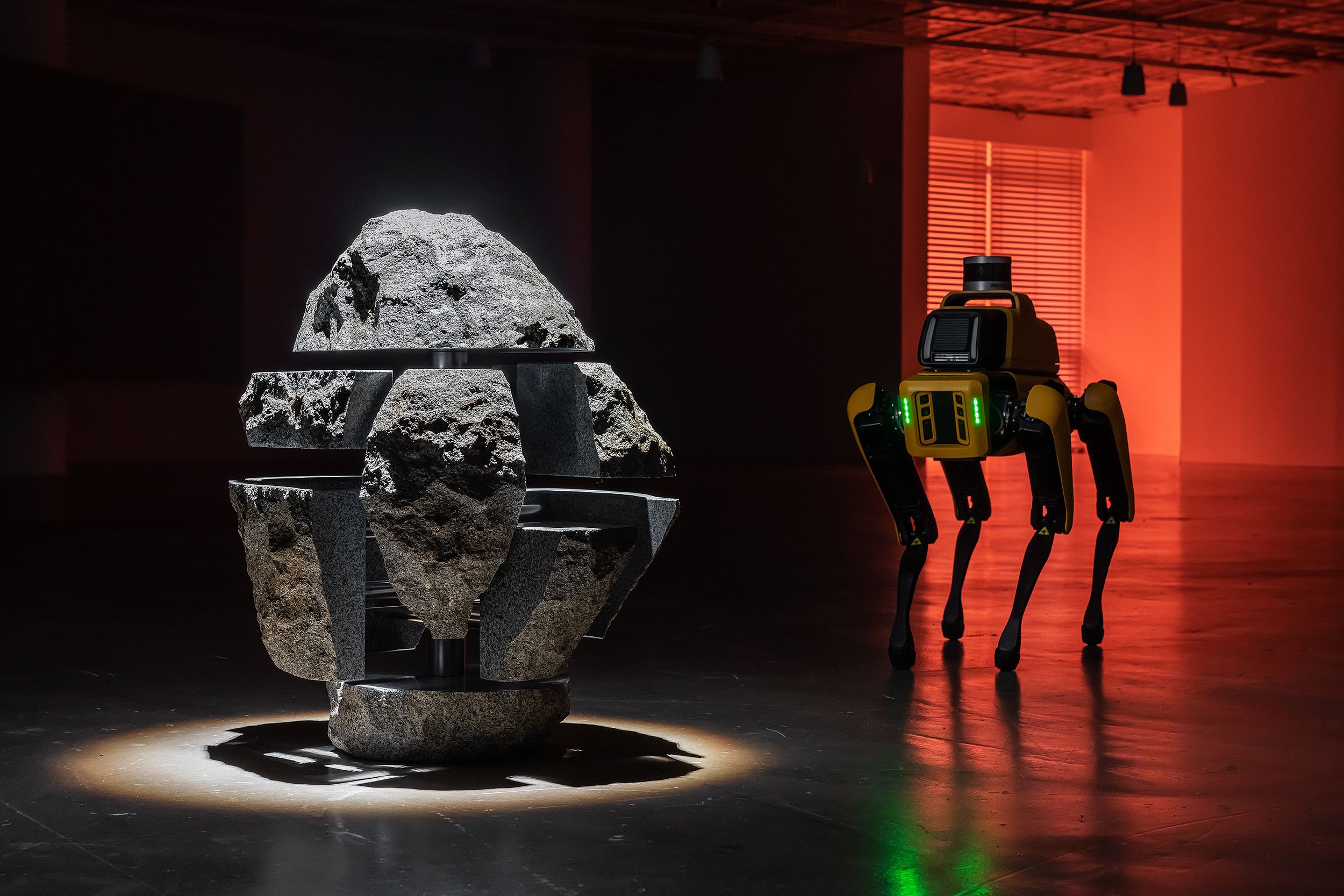A four-legged yellow robot studies a rock puzzle installation, illuminated by a red light in the background.