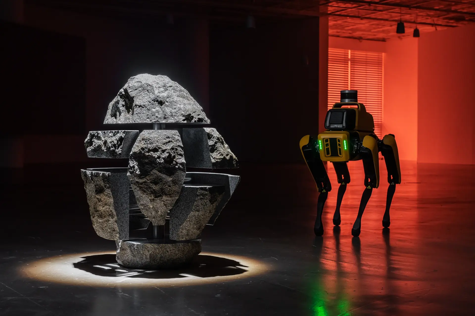 A four-legged yellow robot studies a rock puzzle installation, illuminated by a red light in the background.