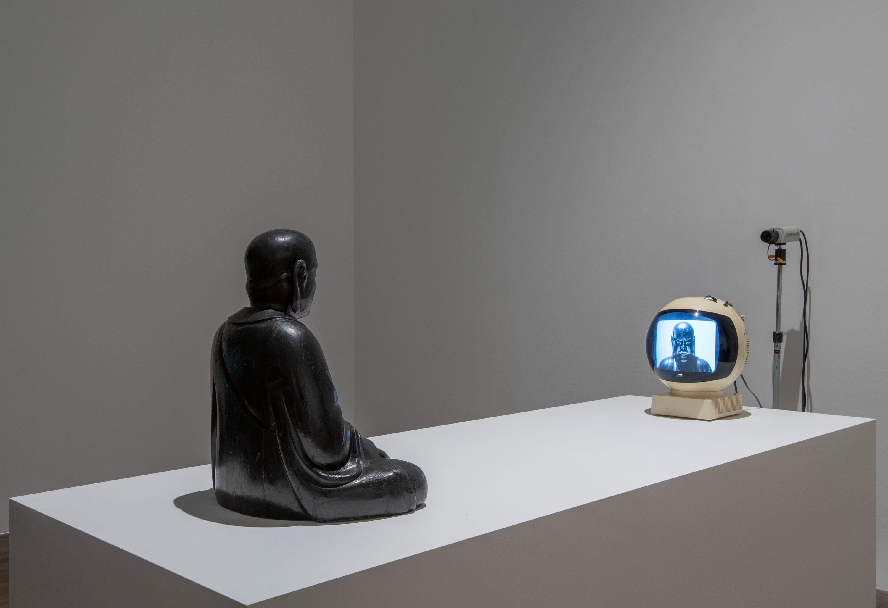 A grey statue of a person is captured in a seated position, engrossed in viewing a small television. The statue is placed ...