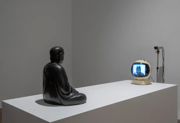 A grey statue of a person is captured in a seated position, engrossed in viewing a small television. The statue is placed ...