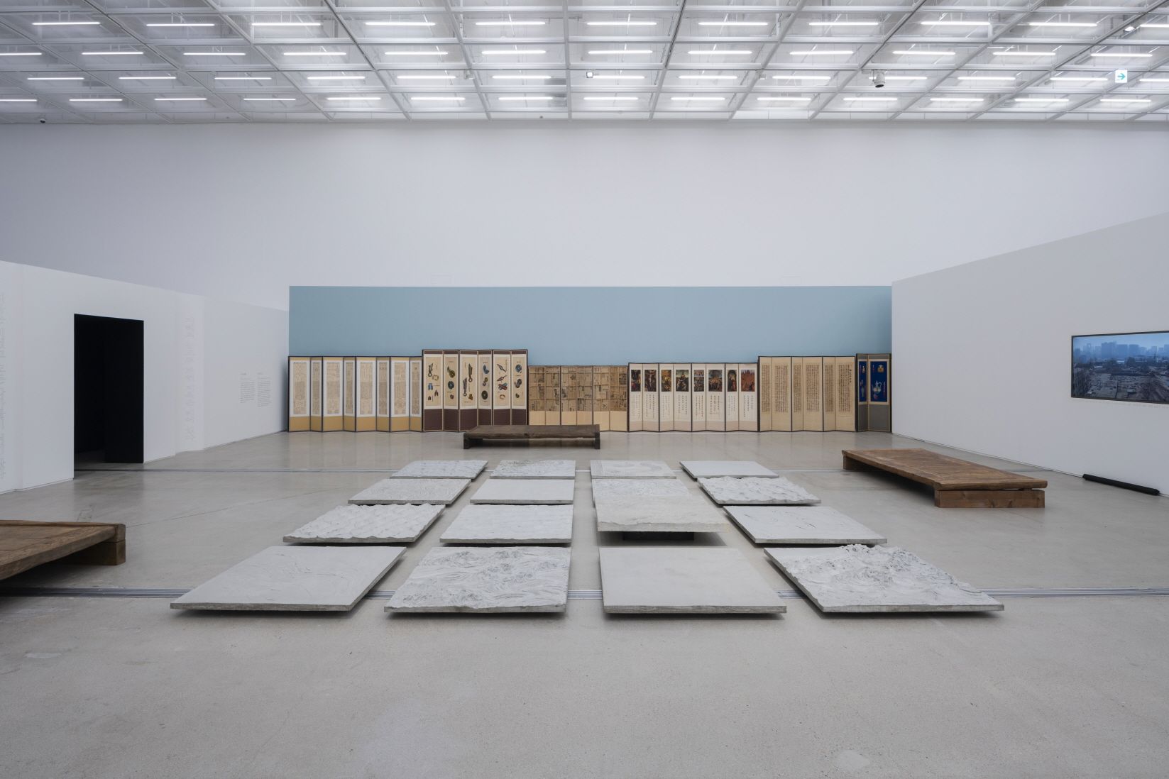 Square objects made of rocky material are arranged in a grid on a gallery floor, with diverse artworks adorning the wall b...
