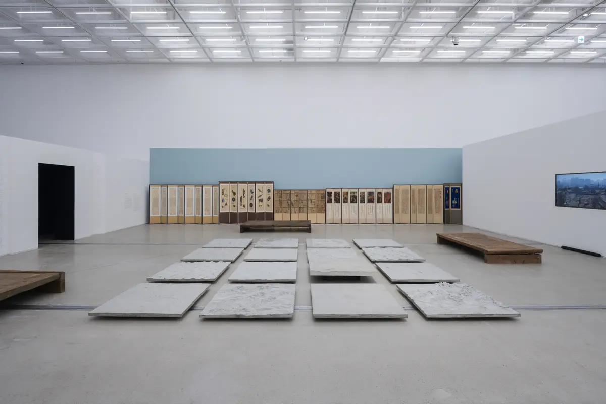 Square objects made of rocky material are arranged in a grid on a gallery floor, with diverse artworks adorning the wall behind.