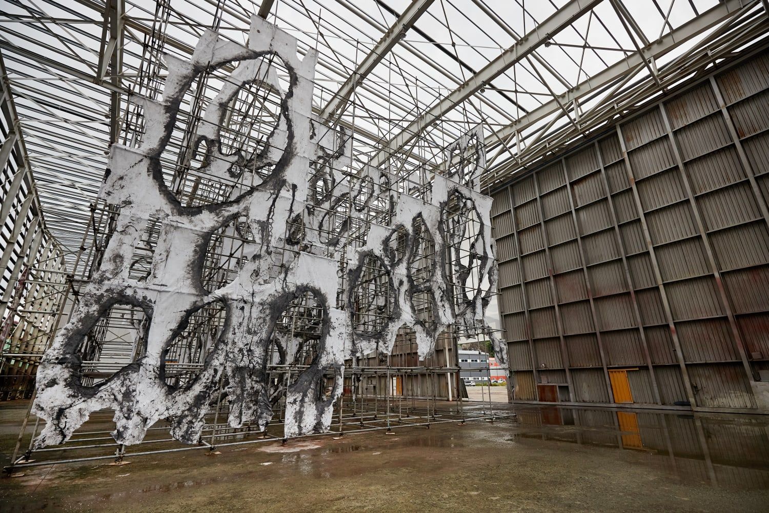 Large outdoor art installation with white and blackened, torn fabric stretched over scaffolding in an industrial space.