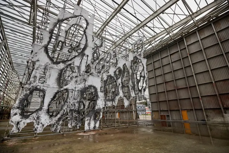Large outdoor art installation with white and blackened, torn fabric stretched over scaffolding in an industrial space.