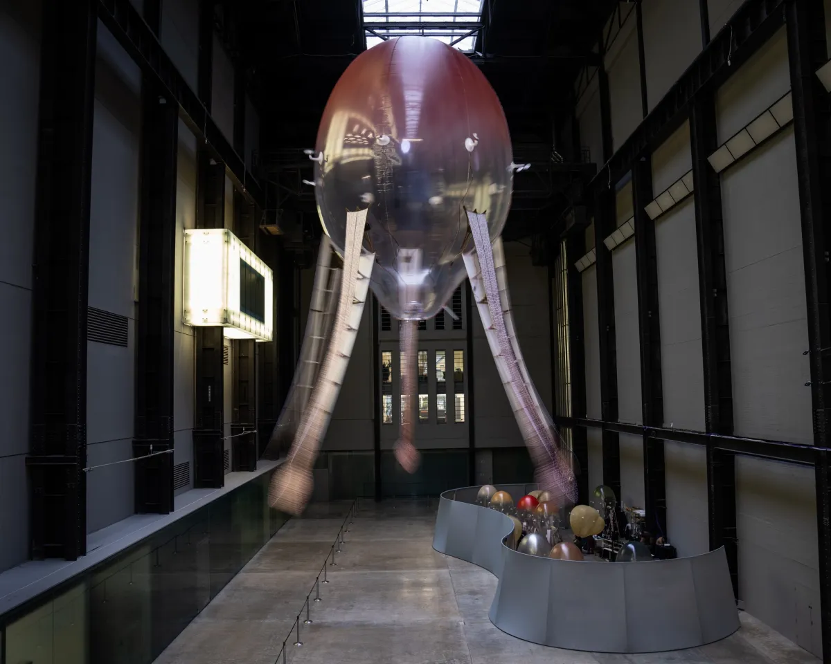 An industrial-styled installation features an octopus-shaped ceiling lamp with snake-headed tentacles, in an expansive gallery hall. Additional lamps appear gathered in a background pool structure.