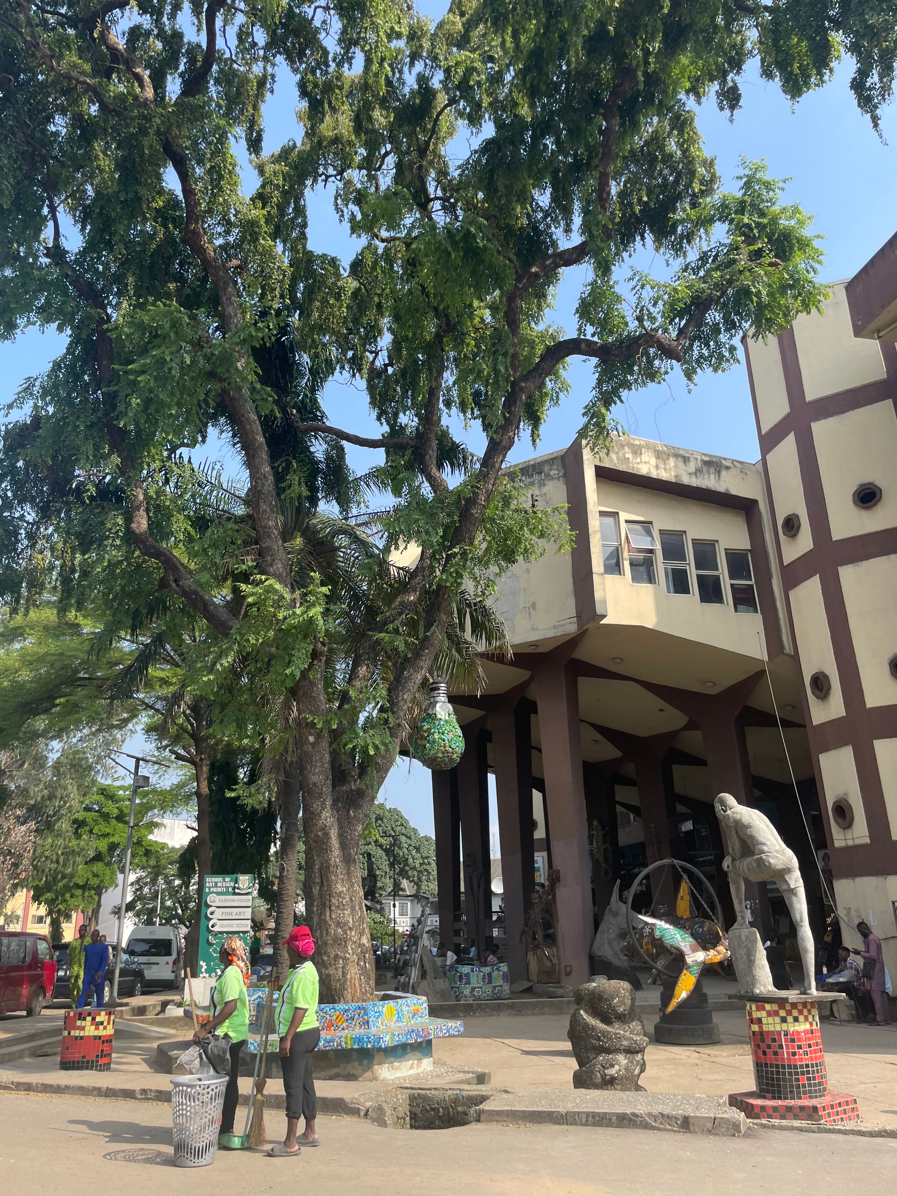 A tree-shaded public square with sculptures and workers cleaning the area.