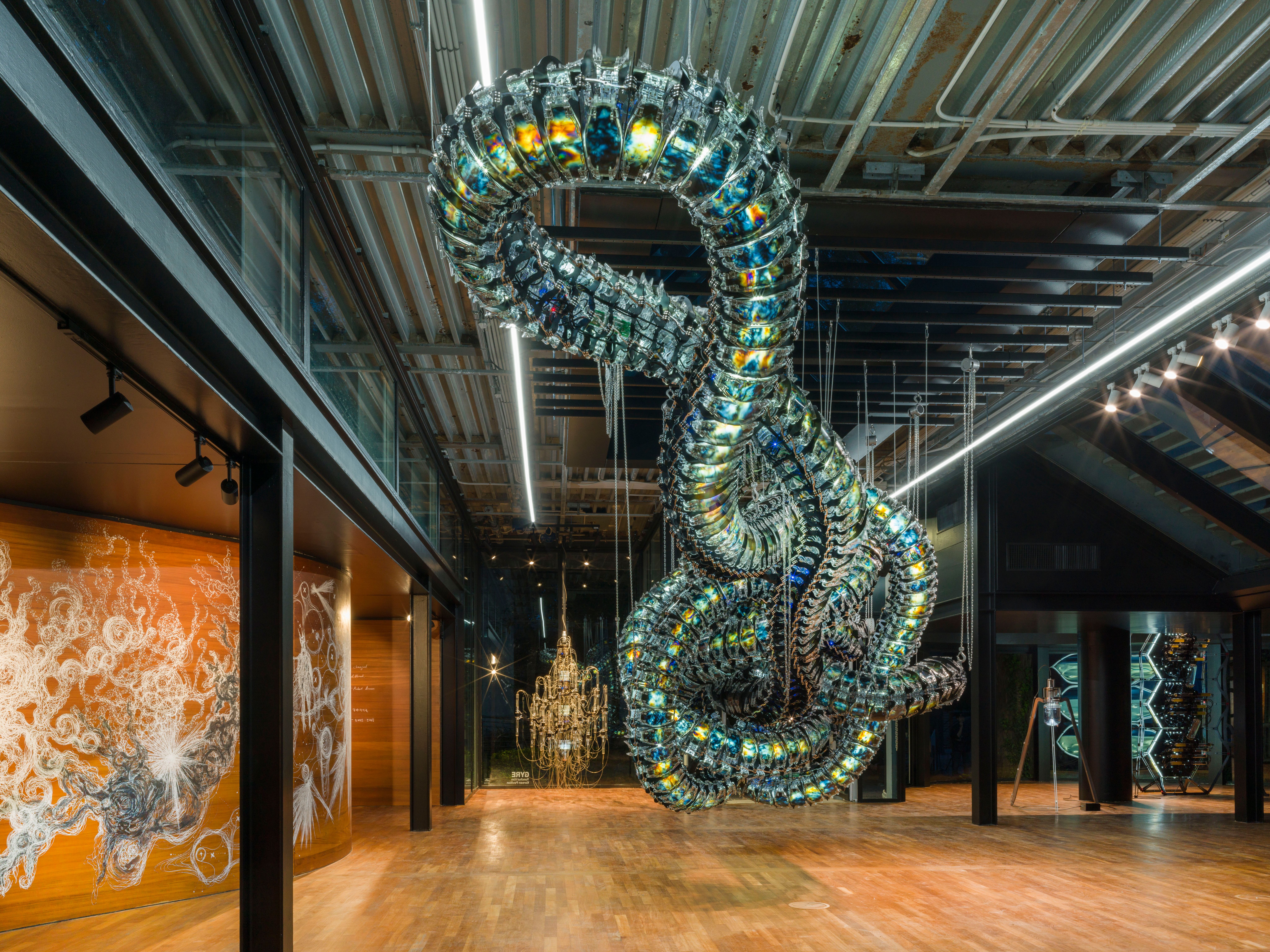 A metallic art installation resembling a snake node is suspended from the ceiling in a gallery hall. Around it, various ot...