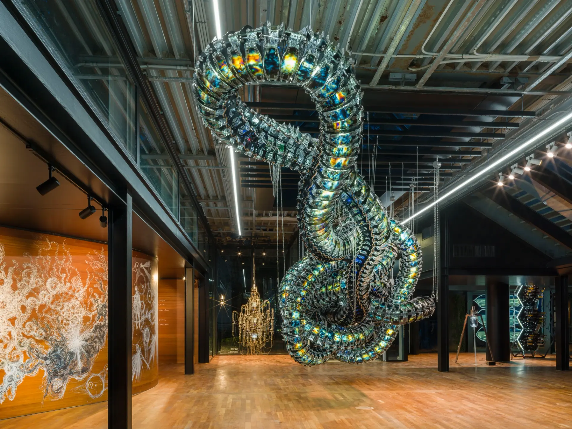 A metallic art installation resembling a snake node is suspended from the ceiling in a gallery hall. Around it, various ot...