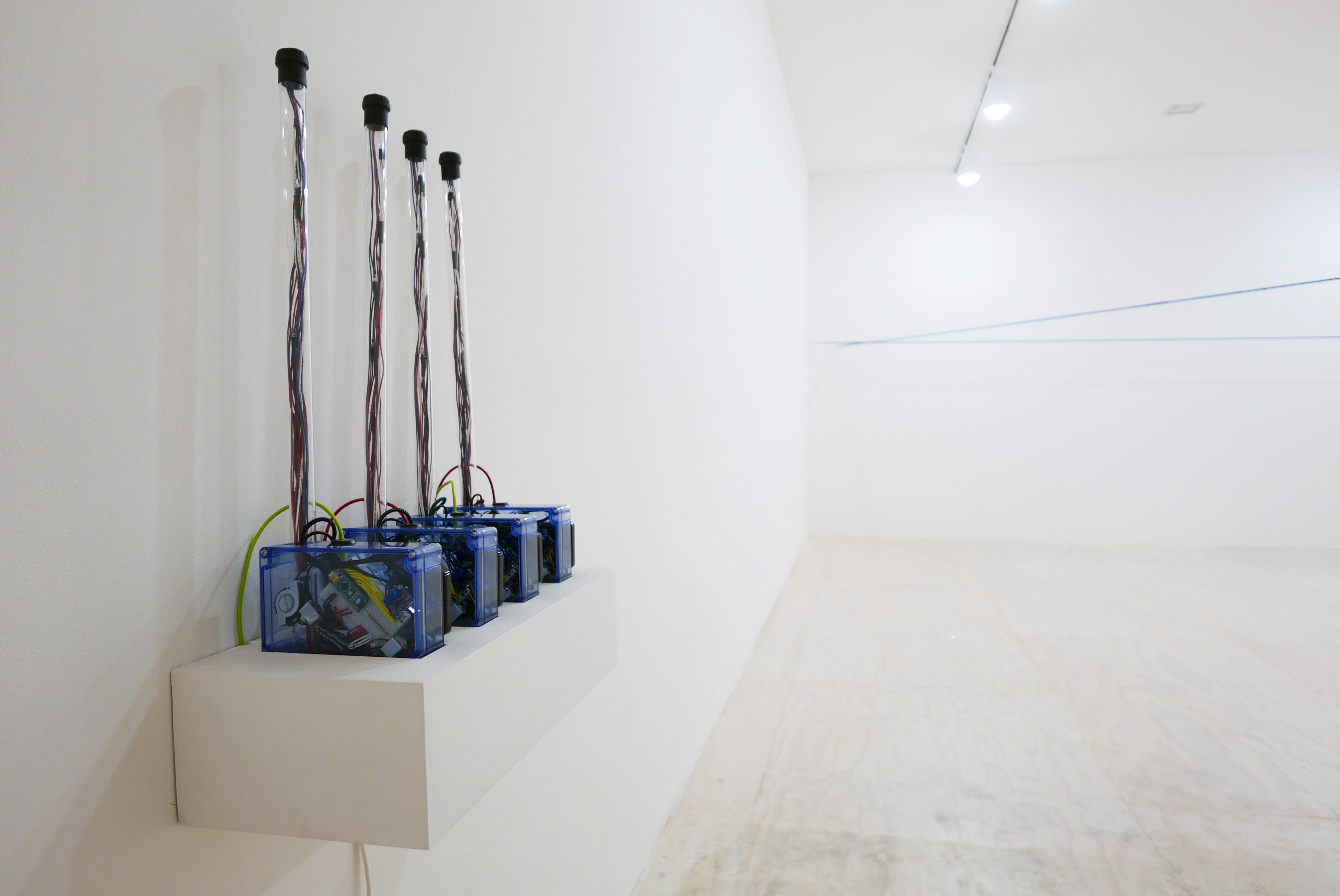 A row of transparent electronic boxes with wires displayed on a shelf in a white-walled gallery.