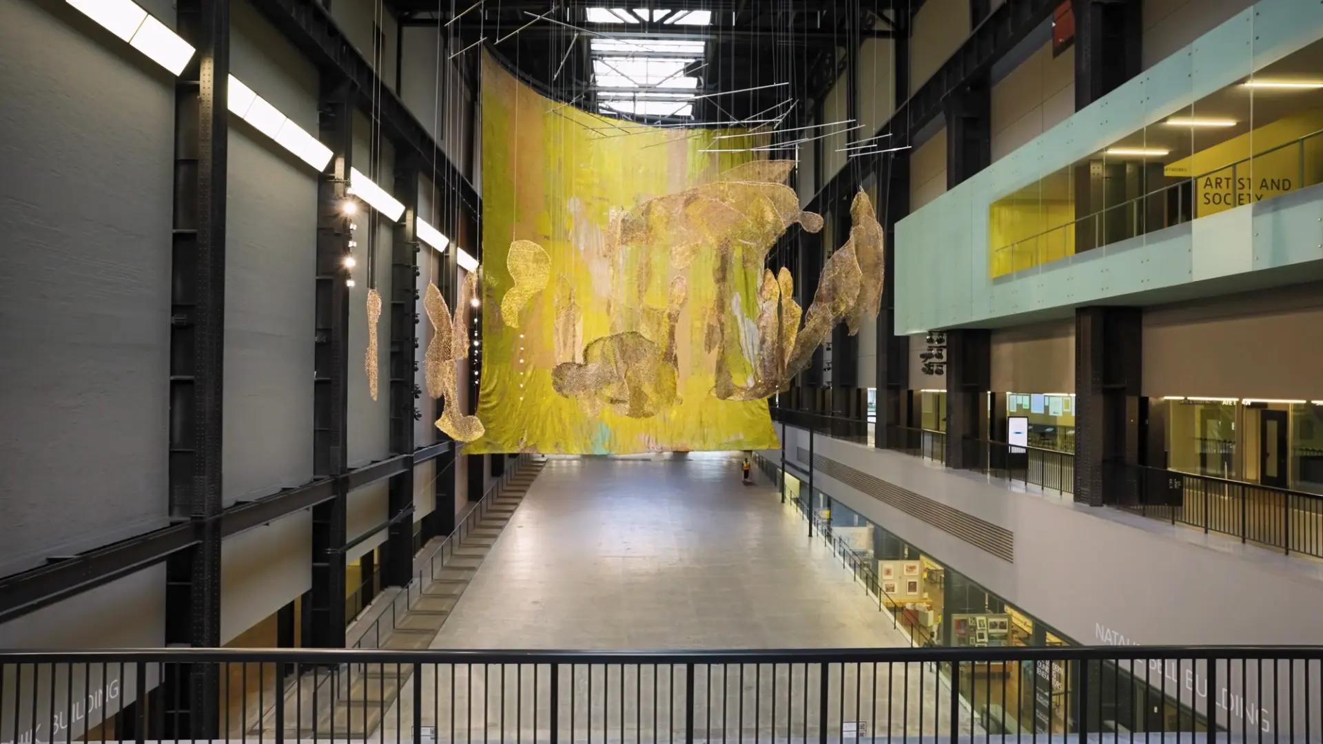 Spacious gallery hall displaying a large yellow art installation with suspended golden figures, evoking a sense of etherea...
