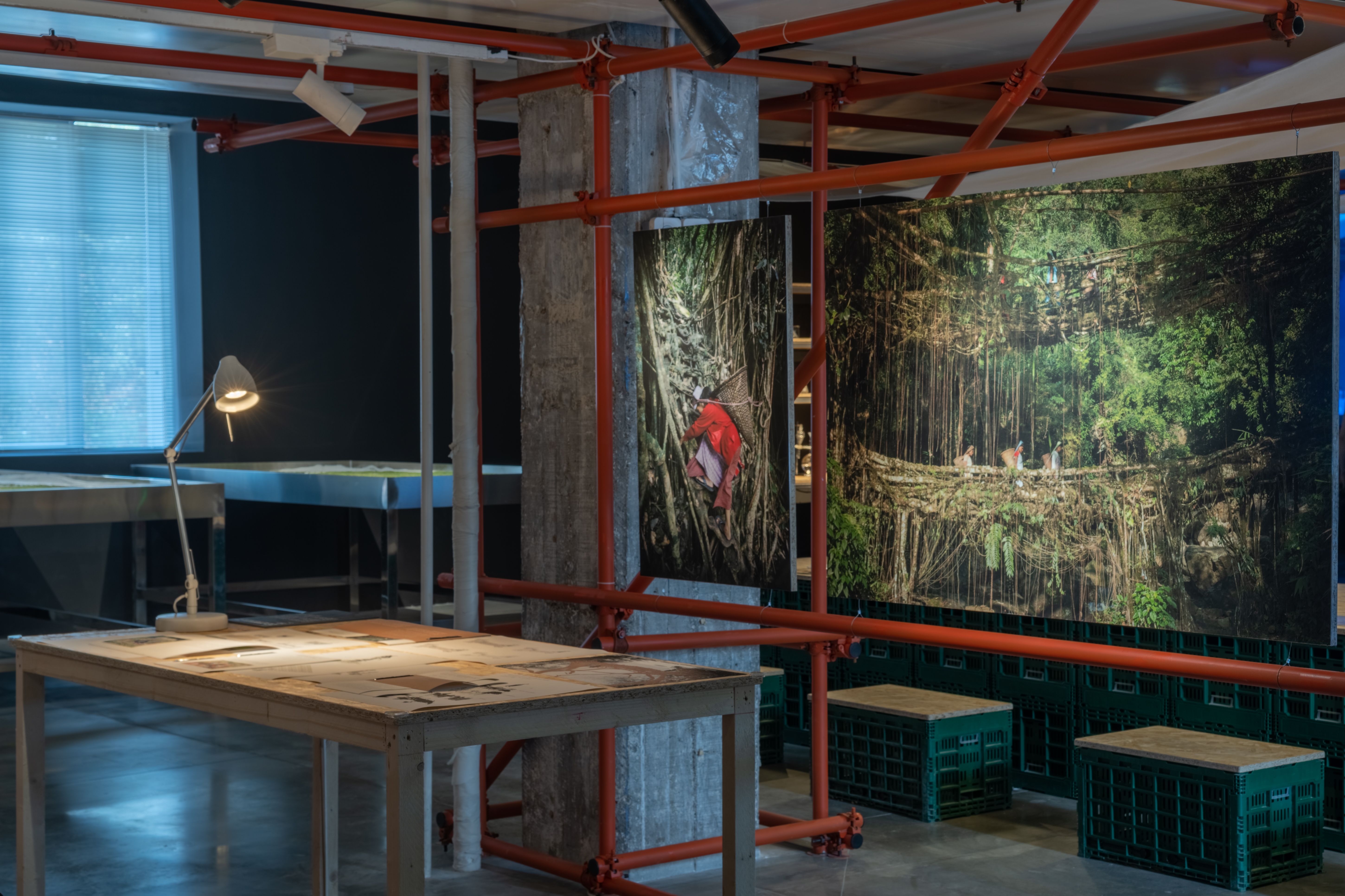 Art space with red scaffolding, photographs, and drawings on a table under a lamp.