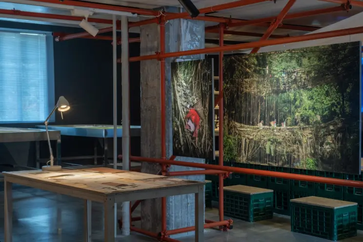 Art space with red scaffolding, photographs, and drawings on a table under a lamp.