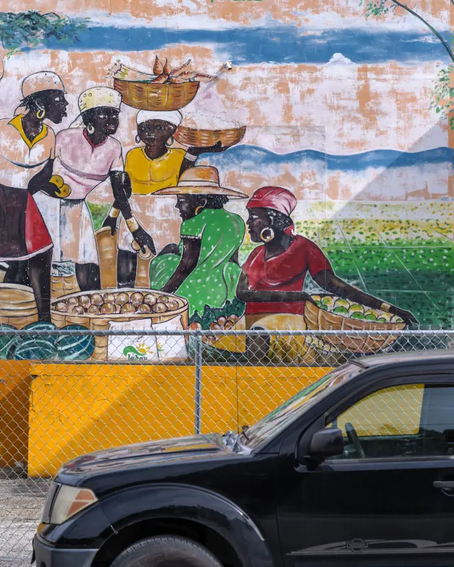 Vibrant mural of women in traditional dress at a market, partially obscured by a parked car.