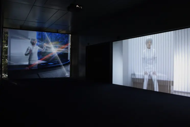 Two projections of a person dressed in white with white hair displayed in an installation. One projection shows the indivi...