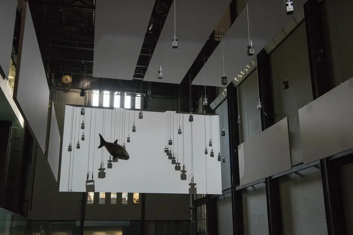 An art installation featuring a fish sculpture suspended from the ceiling in a spacious gallery hall. White panels adorn the surrounding space.