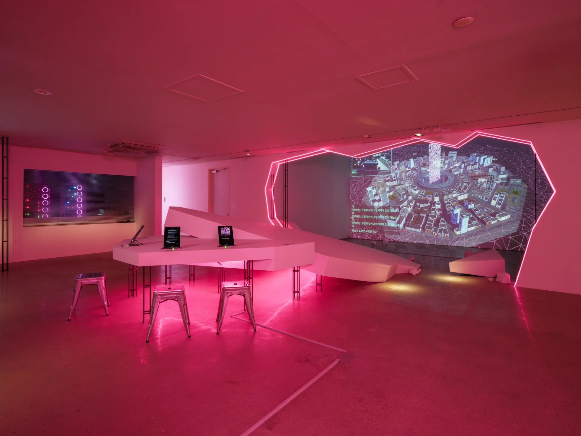 A neon-lit gallery bathed in pink light, featuring a polygonal projection portal displaying a digital cityscape. In the foreground, a faceted white table with stools and mounted tablets evokes a sci-fi command center. On the left, a glass wall reveals circular LEDs arranged in server-like towers.