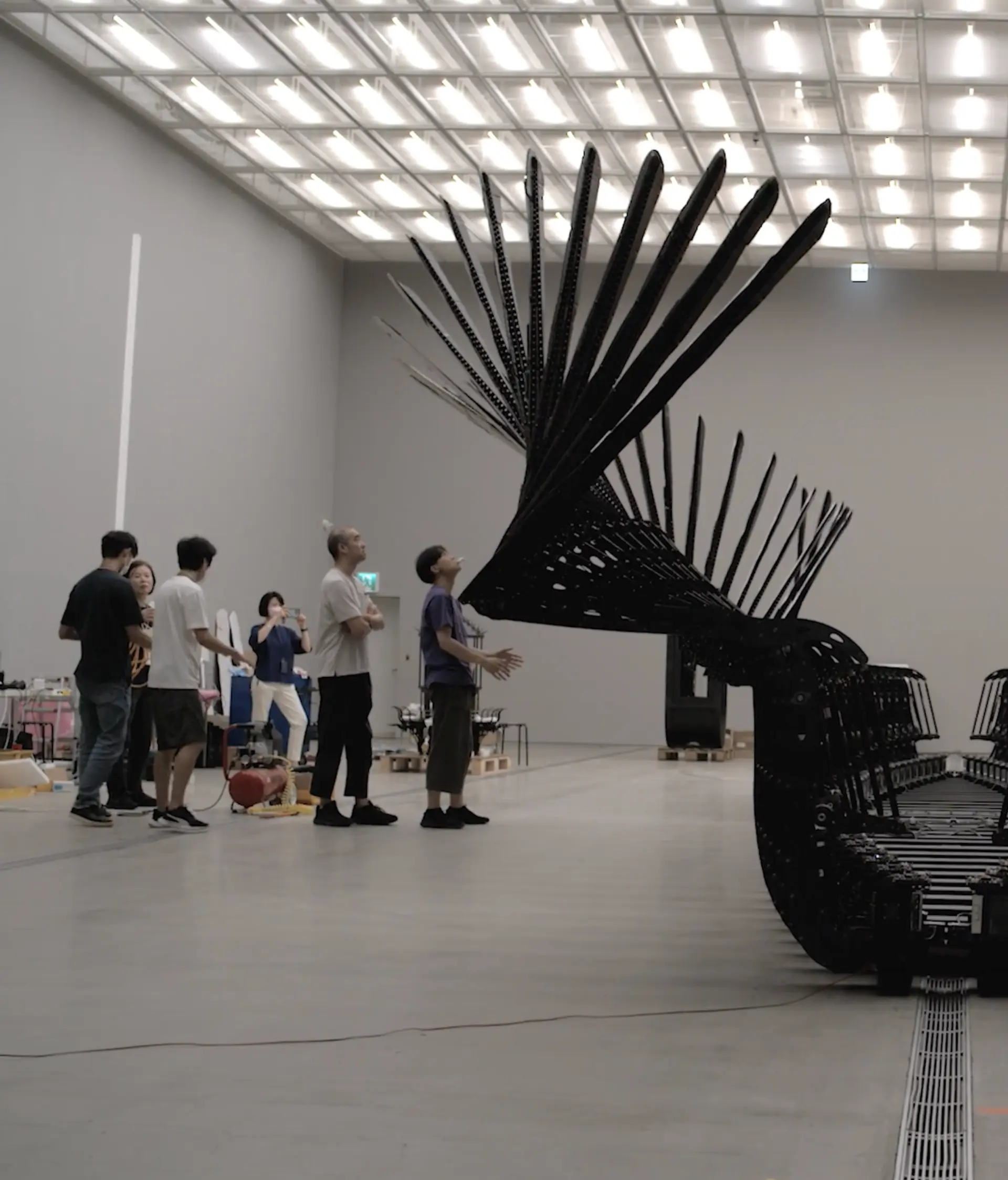 A group of individuals standing in an art gallery, observing a sculpture composed of black, machine-like forms.