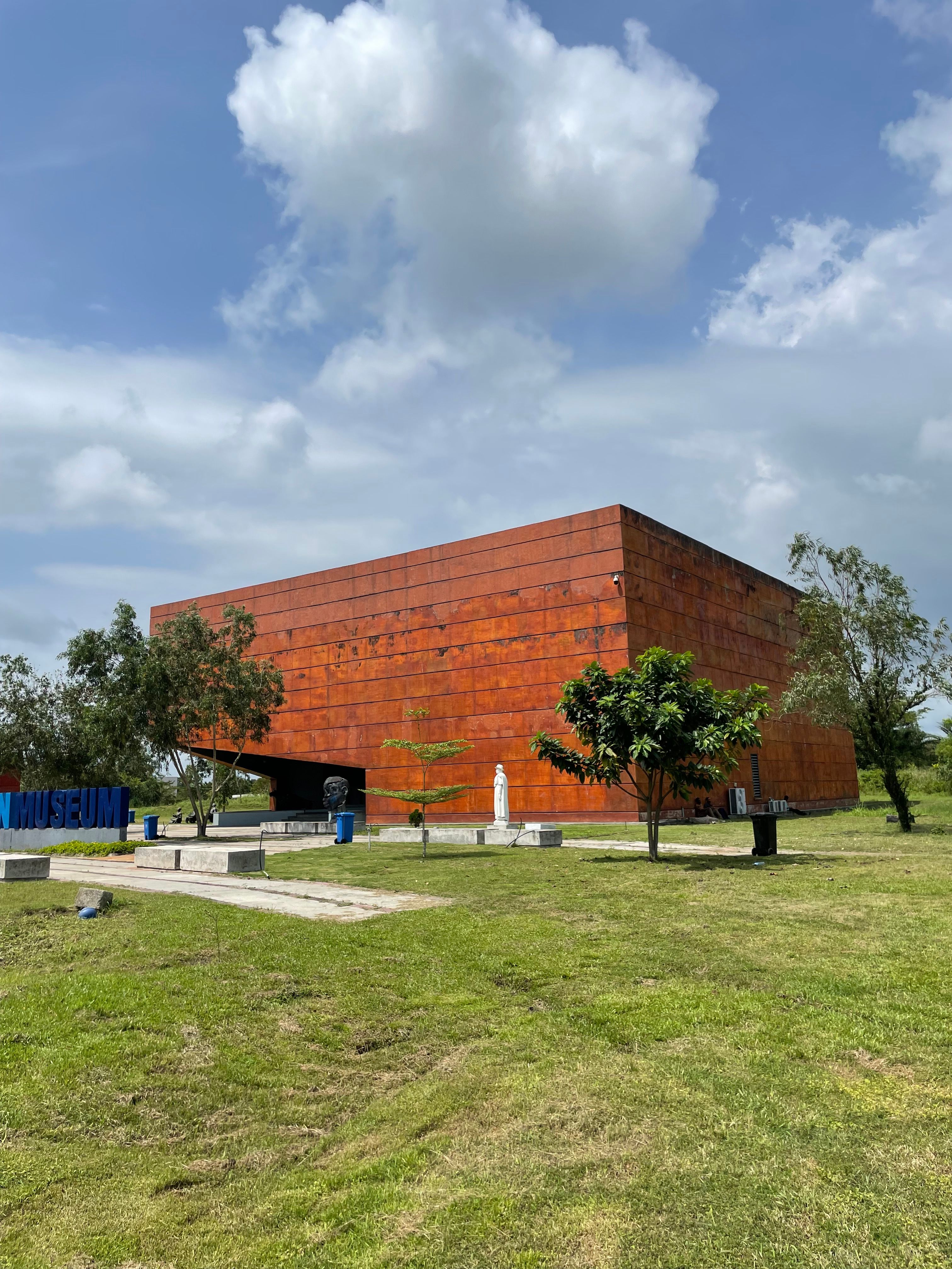 A rust-colored modern museum building surrounded by greenery under a blue sky.