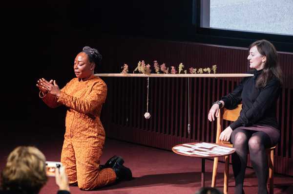 Two people engage on a stage, with a horizontal stick suspended between them, adorned with leaves and various objects.