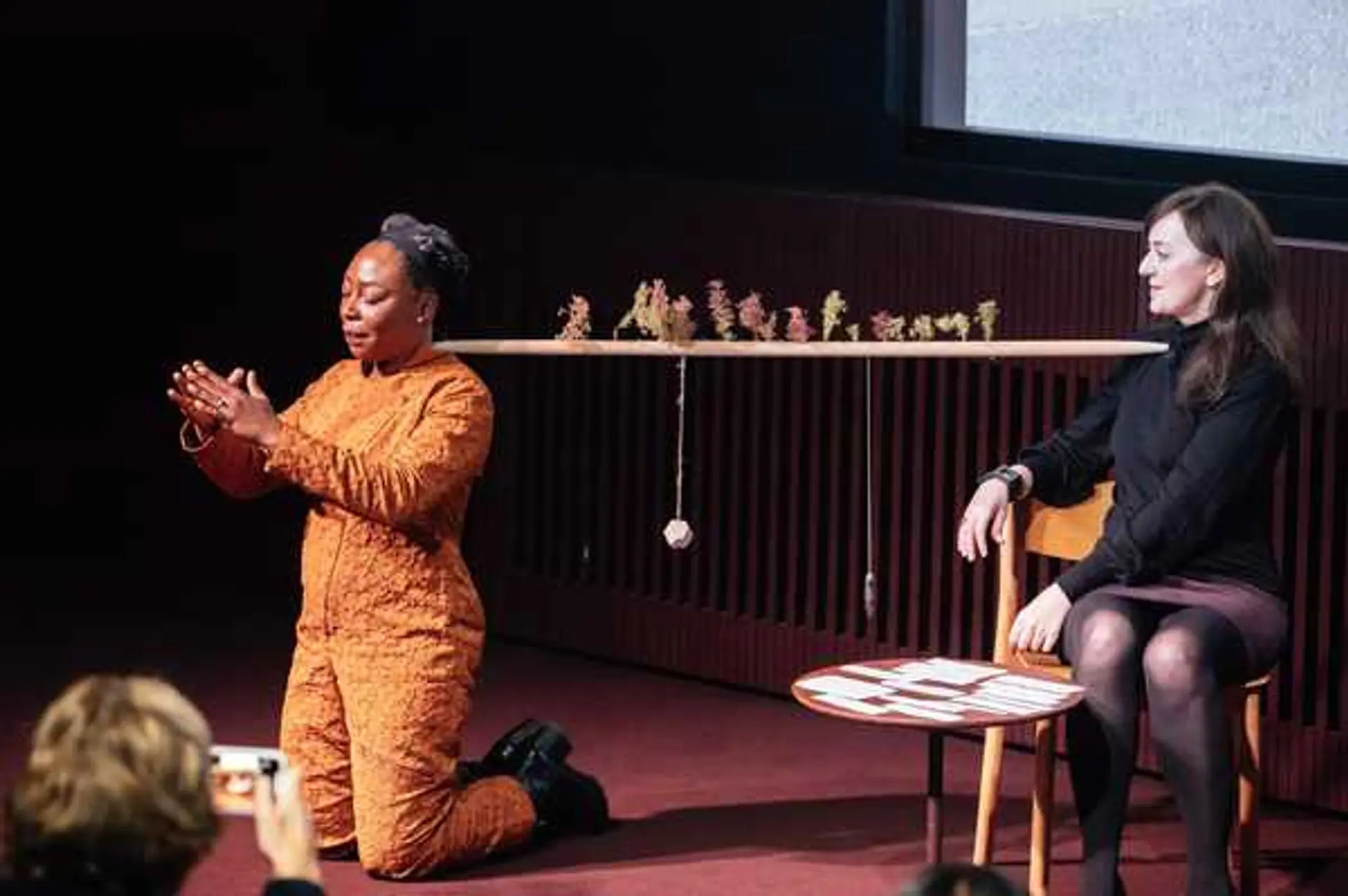 Two people engage on a stage, with a horizontal stick suspended between them, adorned with leaves and various objects.