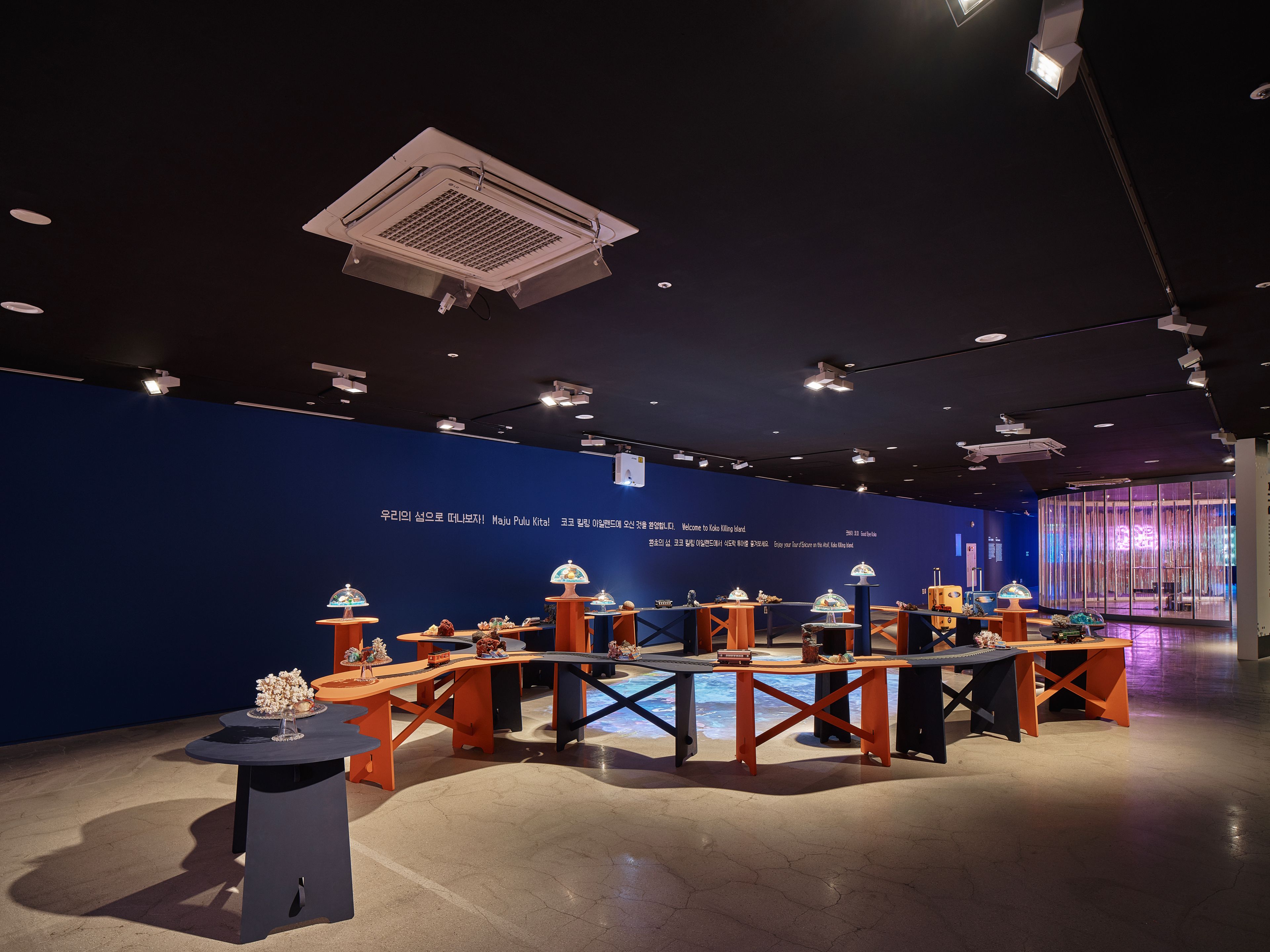 An installation consisting of tables arranged in a circular pattern, forming an atoll-like shape. The tables are set for a...
