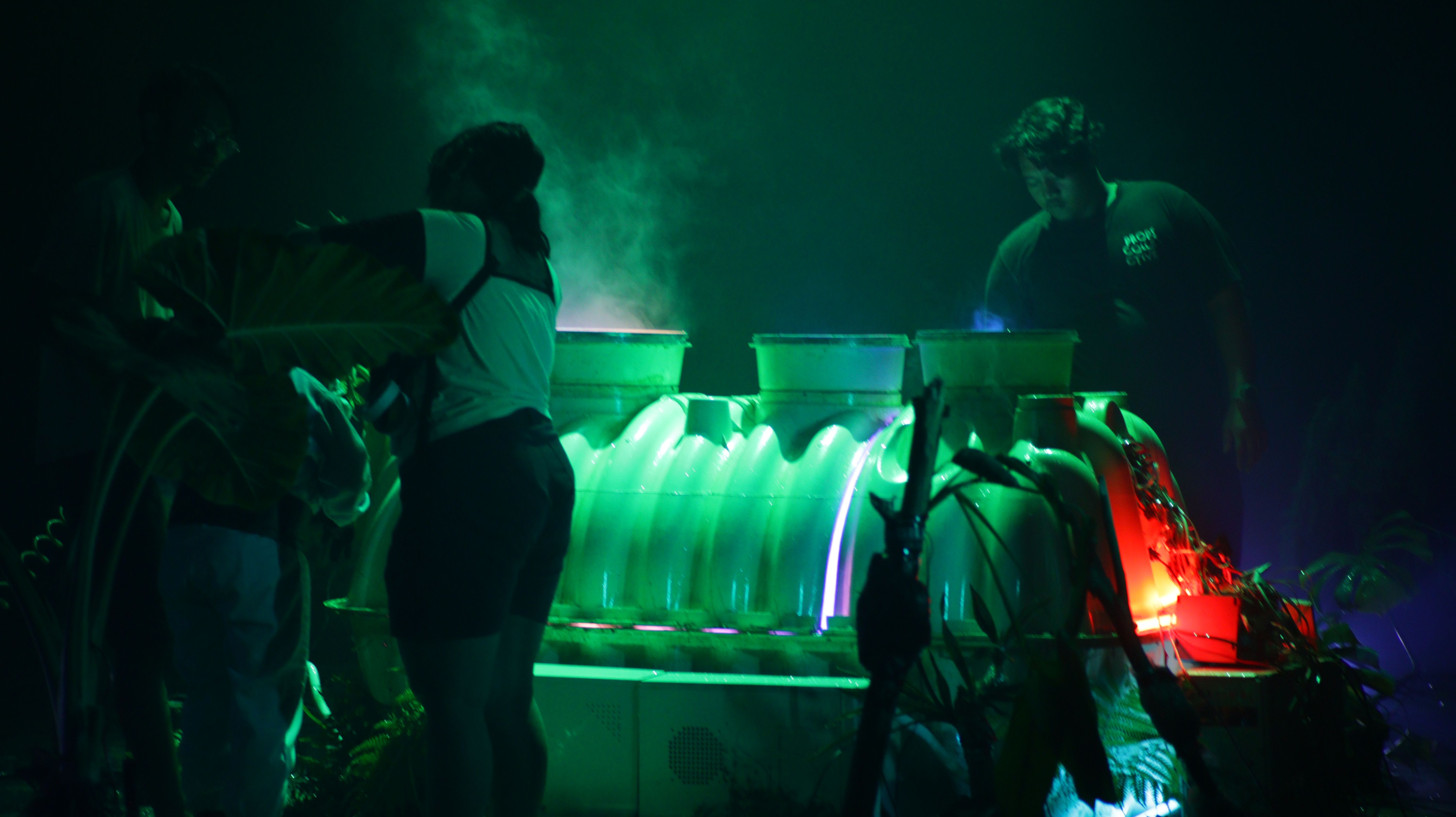 Several individuals are working on a large installation under a glowing green light. The environment is hazy with smoke.