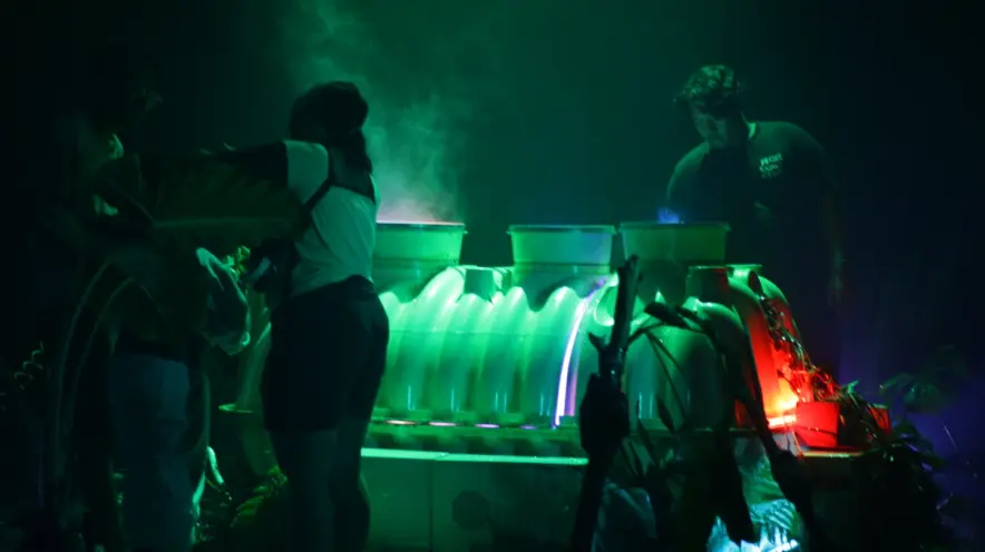 Several individuals are working on a large installation under a glowing green light. The environment is hazy with smoke.