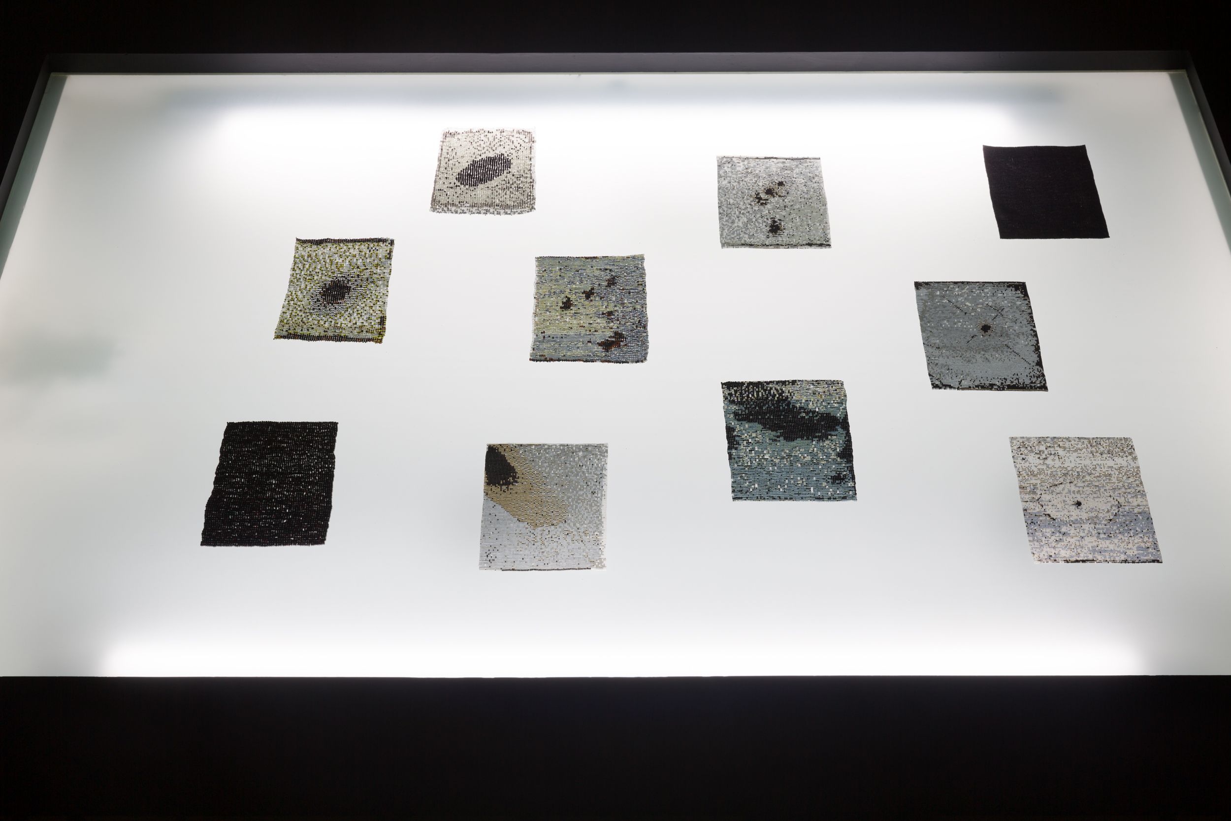 Ten rectangular glass bead tapestries, each sized at 4 by 6 inches, are arranged on a luminescent white LED panel. The intricate designs on the tapestries gleam under the bright light.