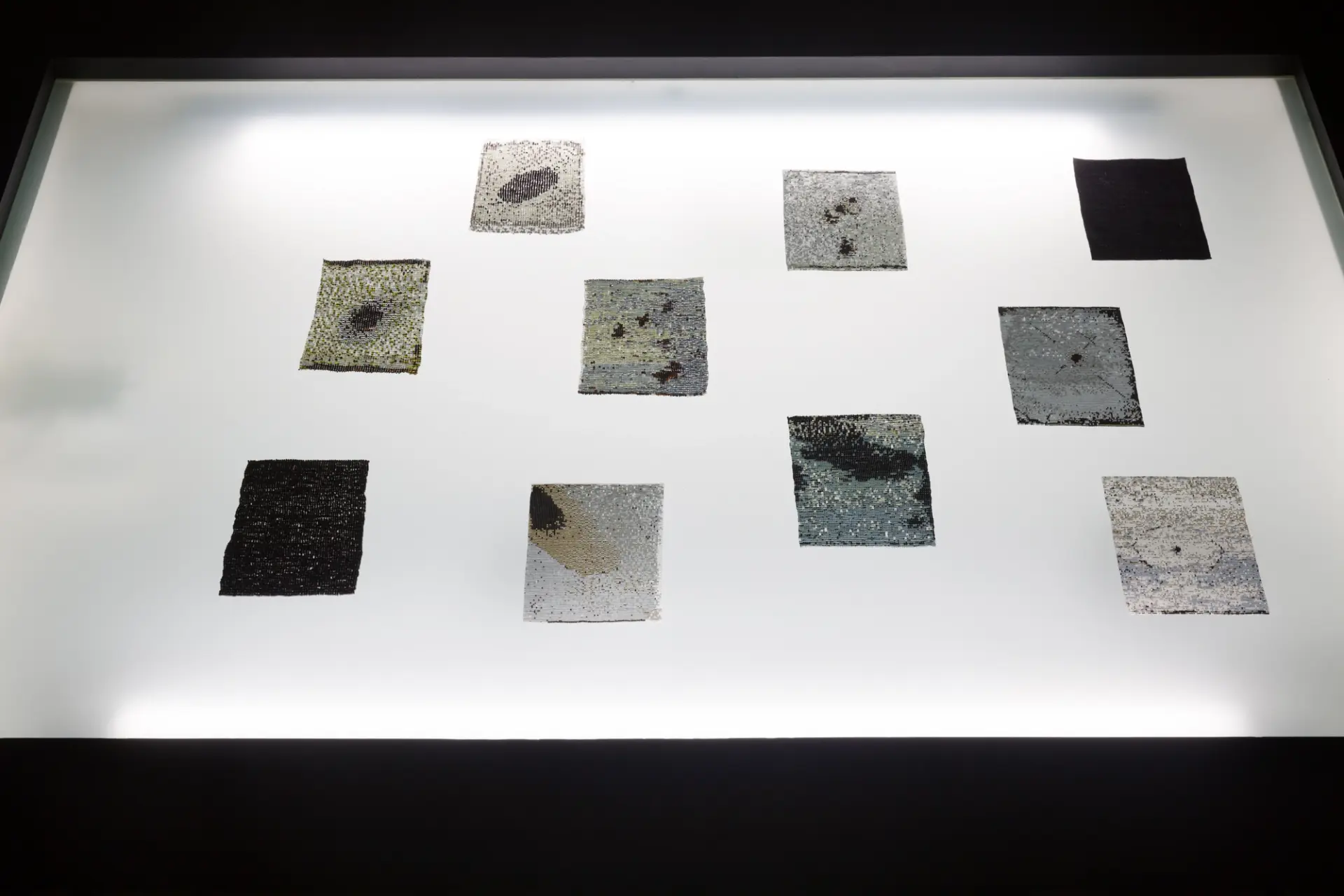 Ten rectangular glass bead tapestries, each sized at 4 by 6 inches, are arranged on a luminescent white LED panel. The intricate designs on the tapestries gleam under the bright light.