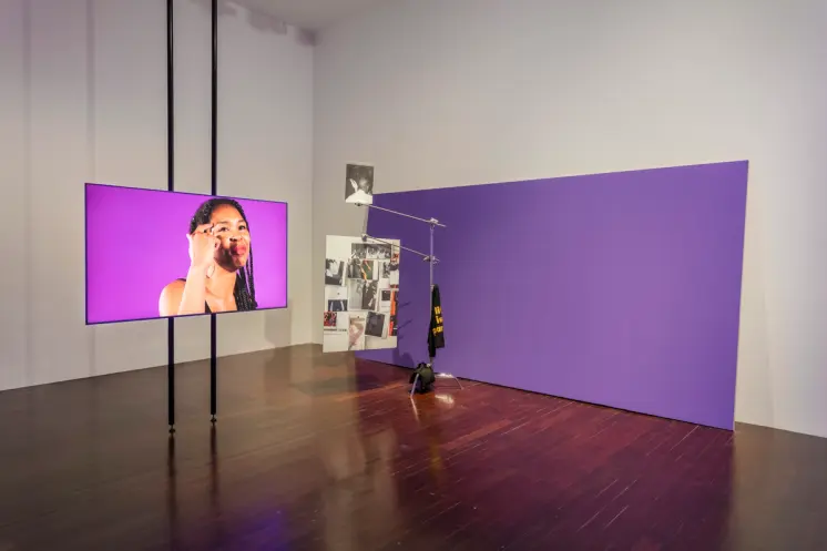 Art installation featuring a large purple rectangle, a photo panel, and a screen displaying an individual against a purple...
