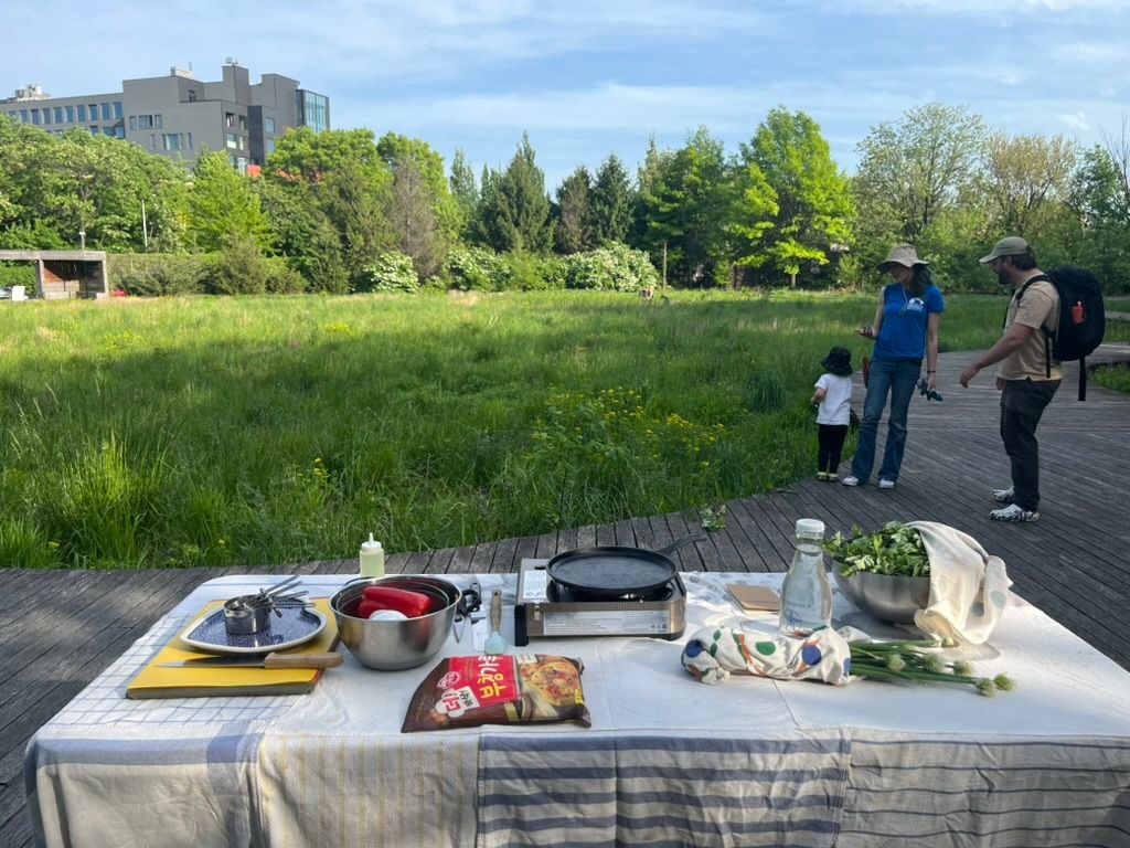A table set up with cooking supplies and ingredients on a wooden deck in a park, with a family in the background near a grassy area.