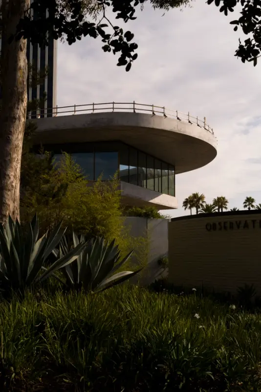 A modern, circular observatory building with large windows and a rooftop railing, surrounded by lush plants and trees under a cloudy sky.