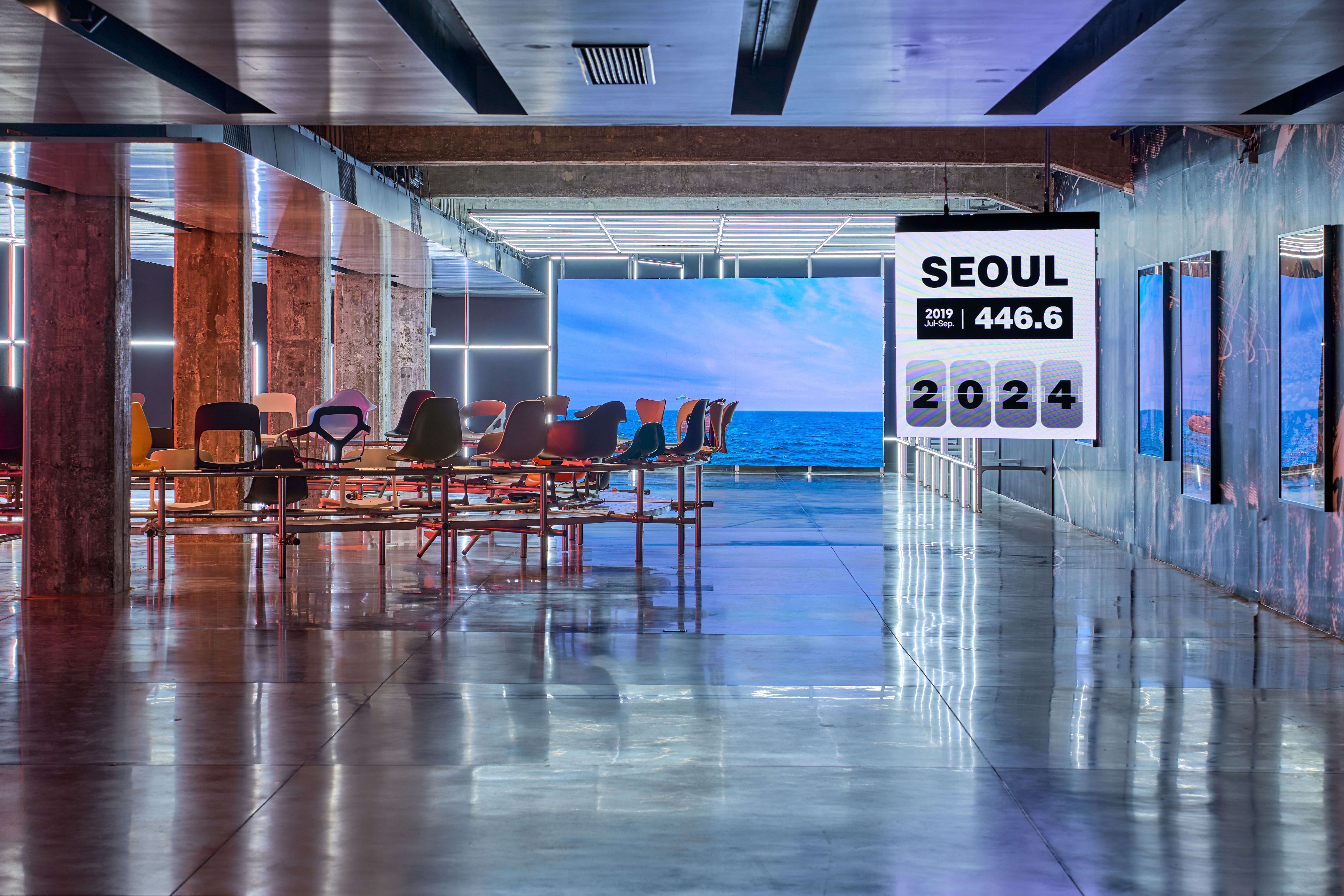 An installation with colorful chairs arranged in a circular pattern within an industrial-style space, featuring large scre...