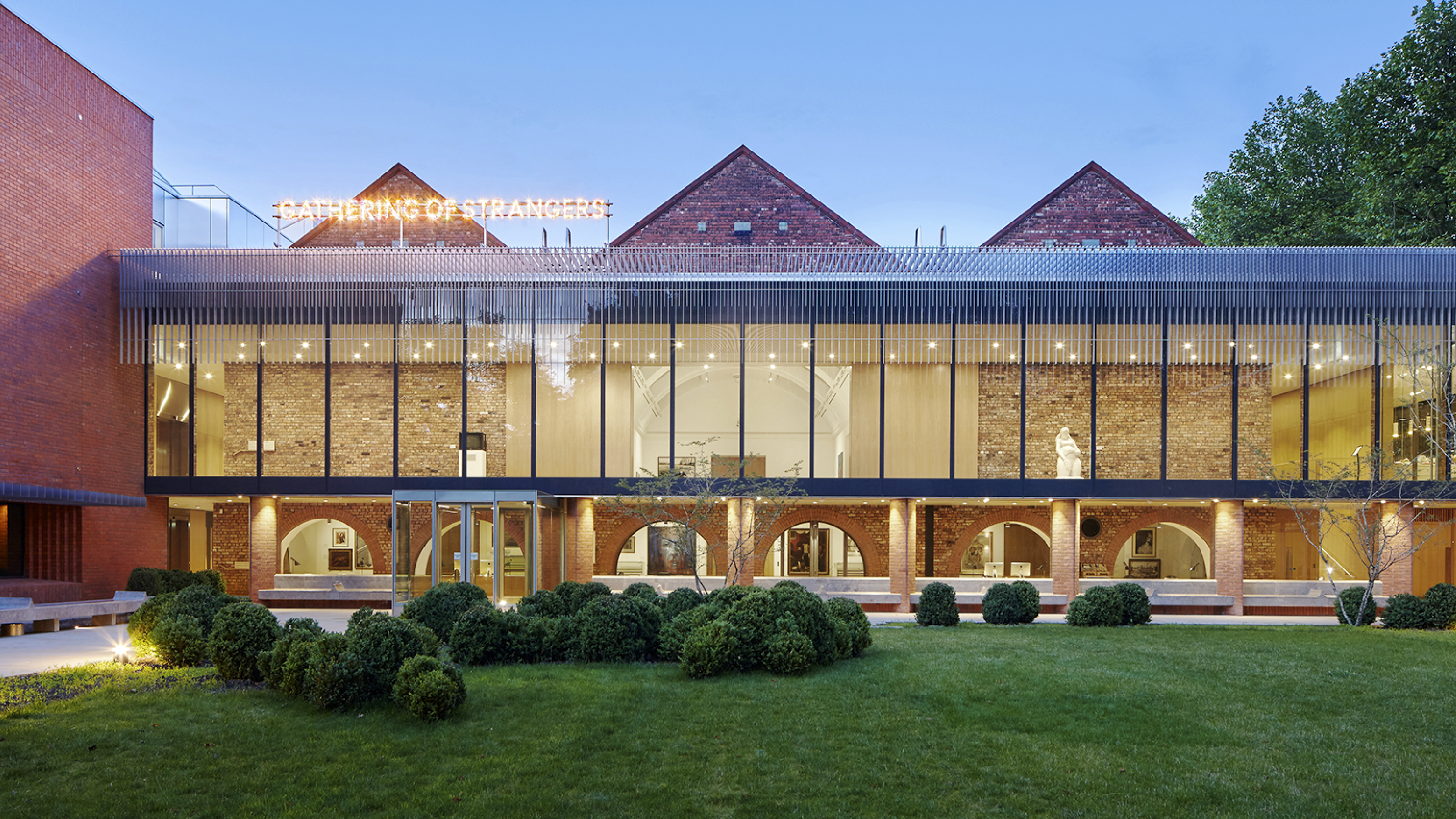 Exterior of the Whitworth gallery at the University of Manchester, showcasing a modern glass and brick facade with arched openings on the ground floor and large windows above. A neon sign on the rooftop reads “GATHERING OF STRANGERS.” The foreground features a manicured lawn with neatly trimmed bushes, and the building is warmly lit against a dusky sky.