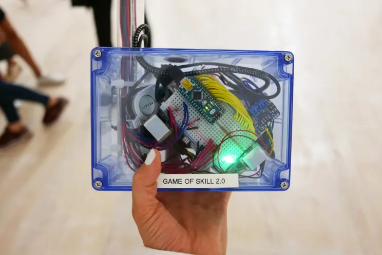 A hand holds a clear blue plastic box labeled “GAME OF SKILL 2.0” filled with wires, a breadboard, and glowing green light.
