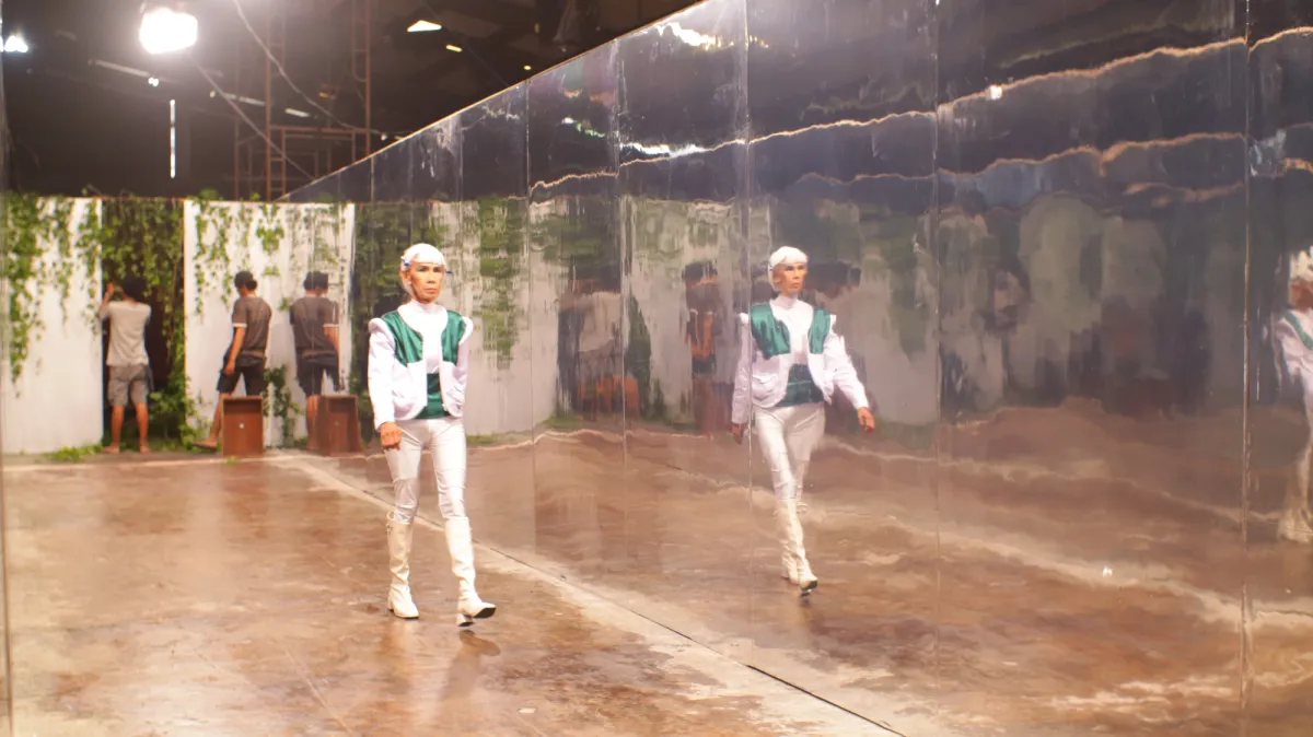 A person in white attire with green details and white hair is walking past a mirrored wall. In the background, two individuals are hanging plants on a white wall.