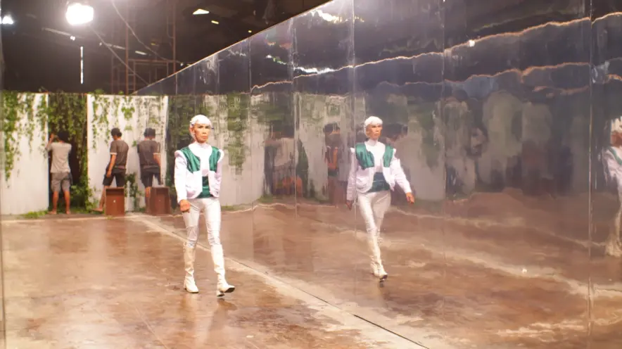 A person in white attire with green details and white hair is walking past a mirrored wall. In the background, two individ...