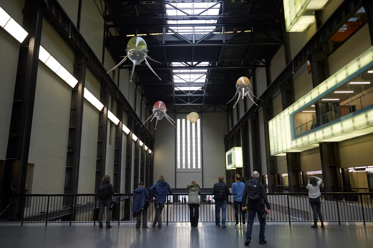 An industrial-style art installation at Tate Modern featuring ceiling lamps shaped like an octopus with snake-headed tentacles. Spectators are scattered around the room, observing the piece against the dimmed background.