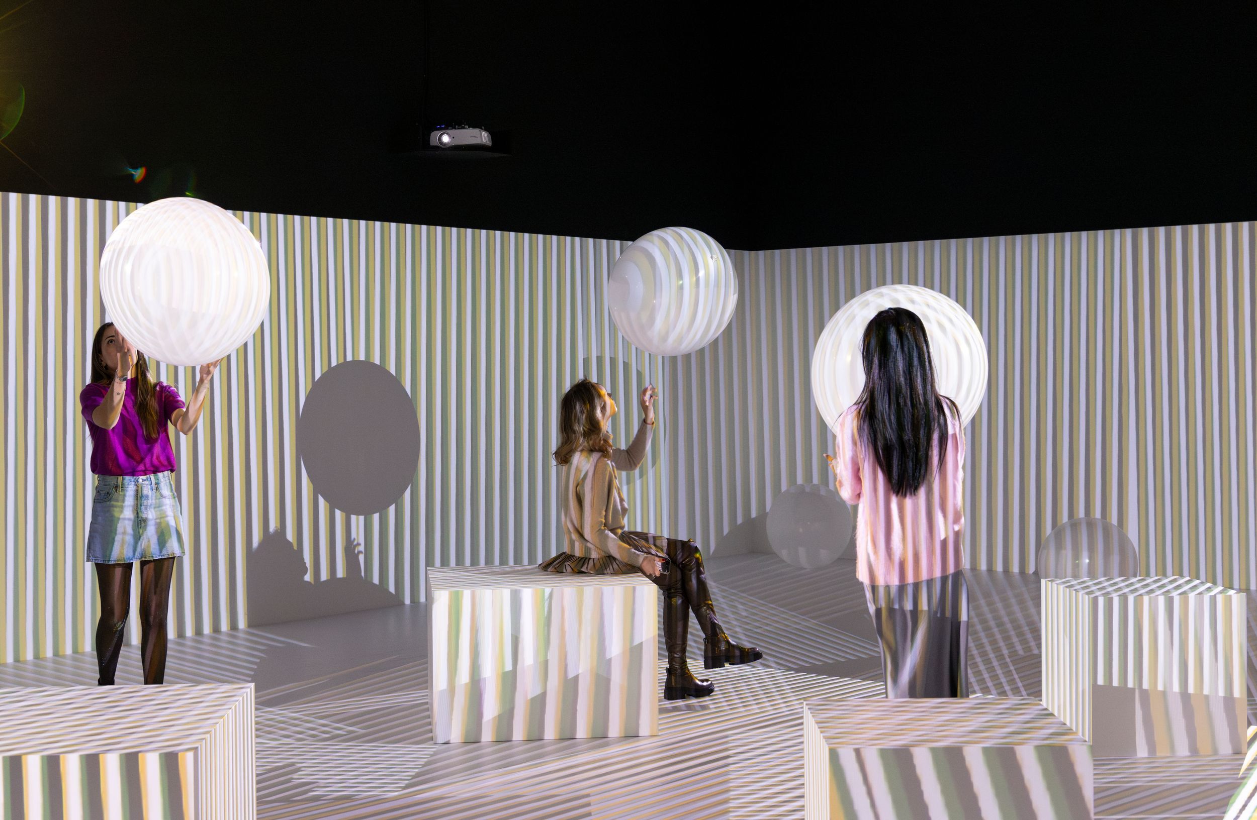 Three women interact with large, glowing spherical objects in an immersive installation space filled with vertical striped...