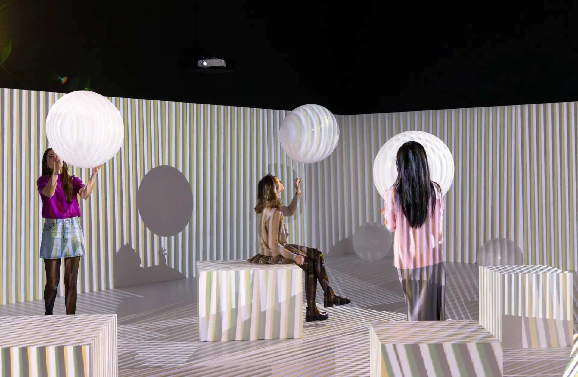 Three women interact with large, glowing spherical objects in an immersive installation space filled with vertical striped light projections. The walls, floor, and cube-shaped seats are covered in multicolored stripes, creating a playful, surreal environment. A ceiling-mounted projector casts the patterns, enhancing the sense of movement and depth.